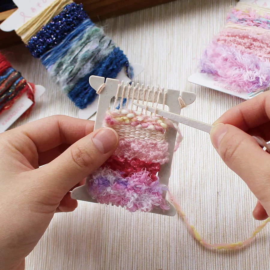 Hands weaving delicate pastel pink, purple, and cream yarns on a Pokeori Pocket Sized Mini Loom with textured embroidery threads and craft supplies in the background.