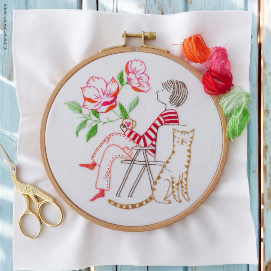 Hand embroidery hoop featuring a whimsical design of a person sitting beside a tiger with colorful flowers, displayed with embroidery floss and gold scissors from the Me & My Tiger Embroidery Kit.