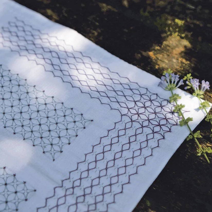 A white fabric textile with a black geometric lattice pattern, laid outdoors on a dark surface with dappled sunlight and small purple flowers resting on one corner.