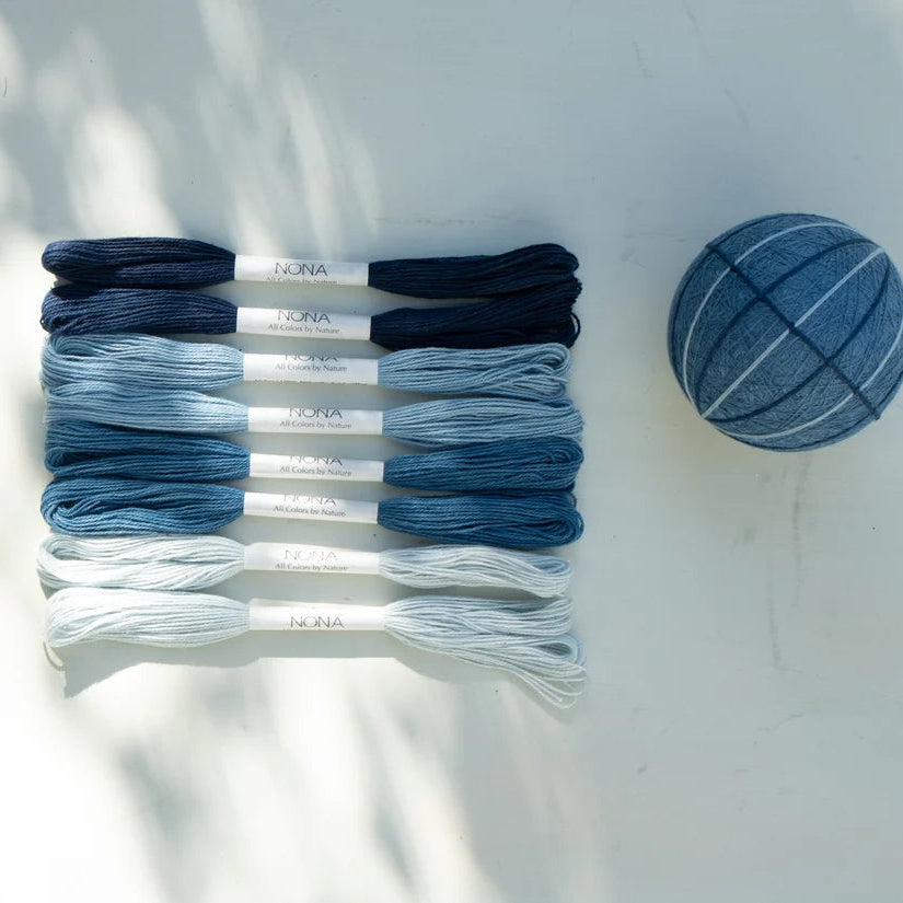 Six skeins of embroidery floss in a blue gradient from dark navy to light icy blue, arranged vertically next to a matching blue striped fabric ball on a white surface with soft leaf shadows.