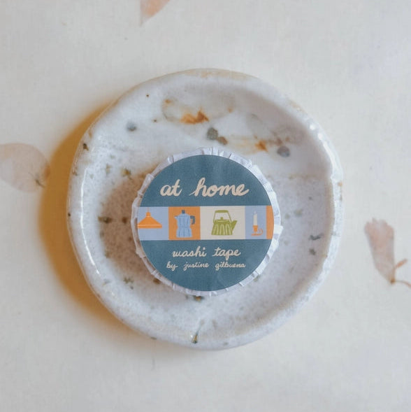 At Home Washi Tape roll with a home-themed design, featuring subtle illustrations on a muted blue background, displayed on a rustic ceramic dish.