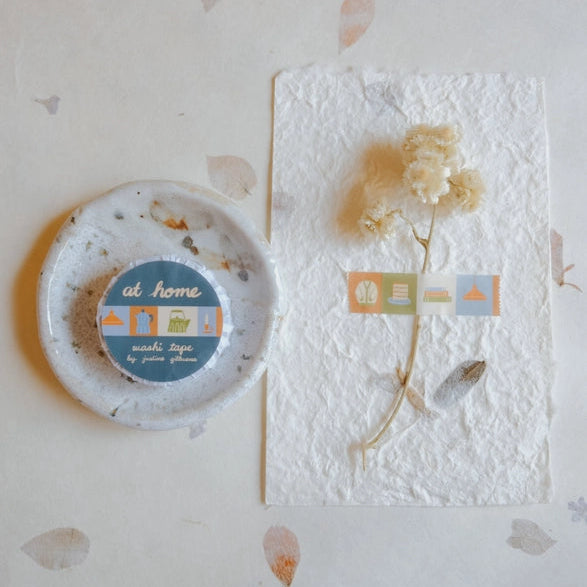At Home Washi Tape with pastel colored house and fabric icons on a textured paper background alongside a small ceramic dish and dried flower, styled for modern needlework crafts.