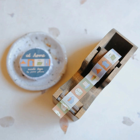 At Home Washi Tape featuring pastel-colored home-themed icons being dispensed from a wooden tape holder, ideal for modern needlework and Japanese stationery crafts.