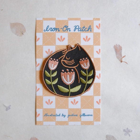 Cat Nap Embroidered Iron-on Patch featuring a black cat curled up among three pink tulip flowers, displayed on a peach and white patterned backing card.