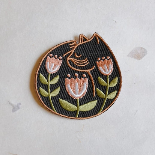 Cat Nap Embroidered Iron-on Patch featuring a black cat curled up with three pink and green flowers, ideal for sewing, quilting, and modern needlework projects.