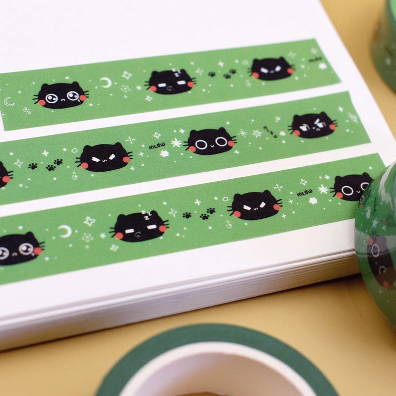 Green washi tape featuring a black cat face pattern, shown partially unrolled on a white sheet with two rolled spools of the same tape on a beige surface.