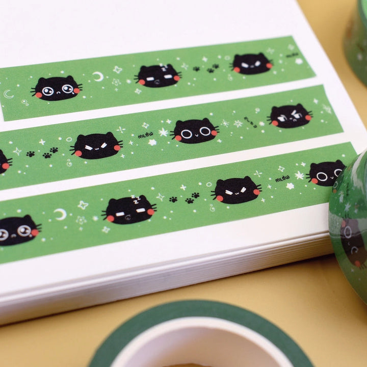 Green washi tape featuring a black cat face pattern, shown partially unrolled on a white sheet with two rolled spools of the same tape on a beige surface.