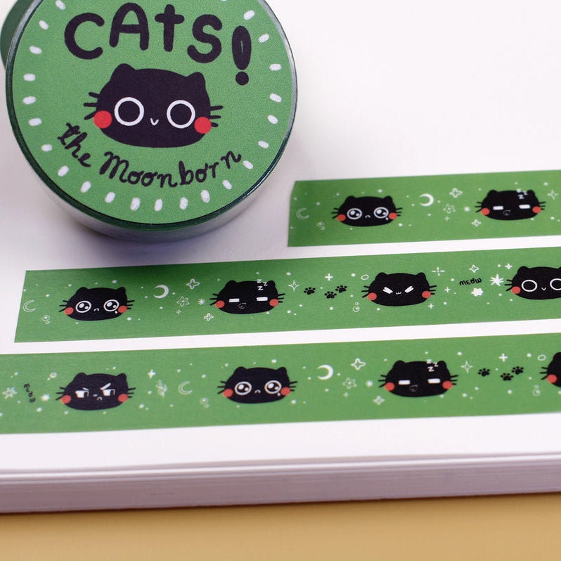Green washi tape with a repeating pattern of black cartoon cat faces, shown unrolled on a white notebook next to its round green packaging label that reads 'CATS! the Moon-born'.