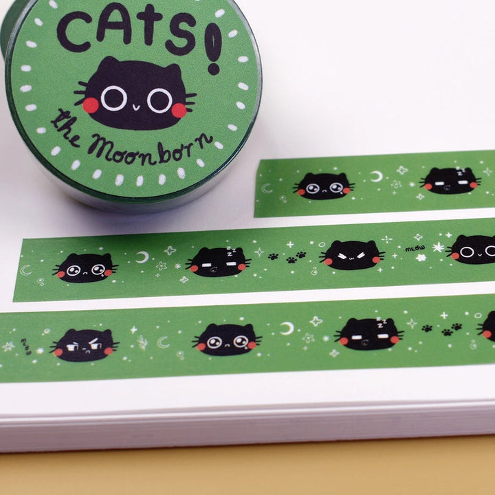 Green washi tape with a repeating pattern of black cartoon cat faces, shown unrolled on a white notebook next to its round green packaging label that reads 'CATS! the Moon-born'.