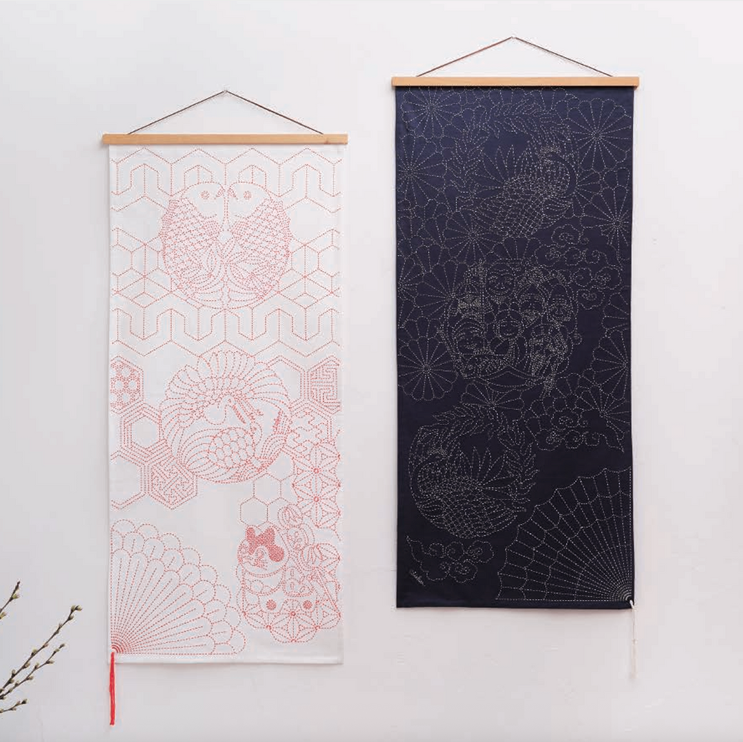 Two vertical hanging fabric wall scrolls with wooden dowels are displayed against a plain white wall. The left scroll is white with pink geometric and fish motifs, and the right scroll is dark navy with light gray floral and fan motifs.
