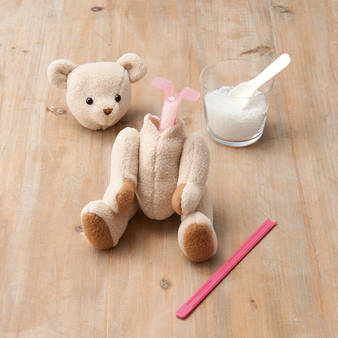 Tube & Plunger Style Stuffing Tool inserted into a partially stuffed plush bear on a wooden surface, accompanied by a small glass of stuffing pellets and a pink plastic clip.