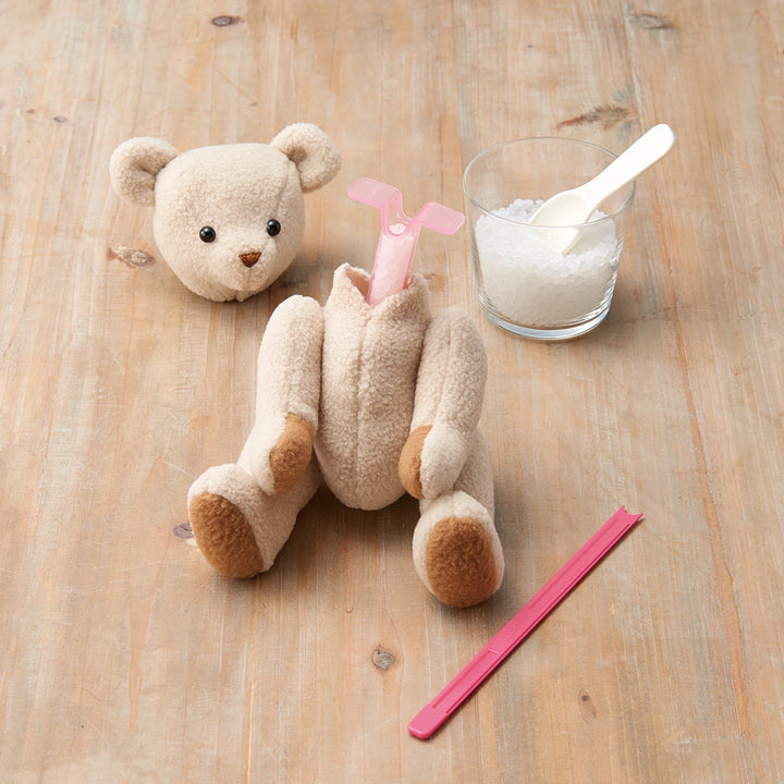 Tube & Plunger Style Stuffing Tool inserted into a partially stuffed plush bear on a wooden surface, accompanied by a small glass of stuffing pellets and a pink plastic clip.
