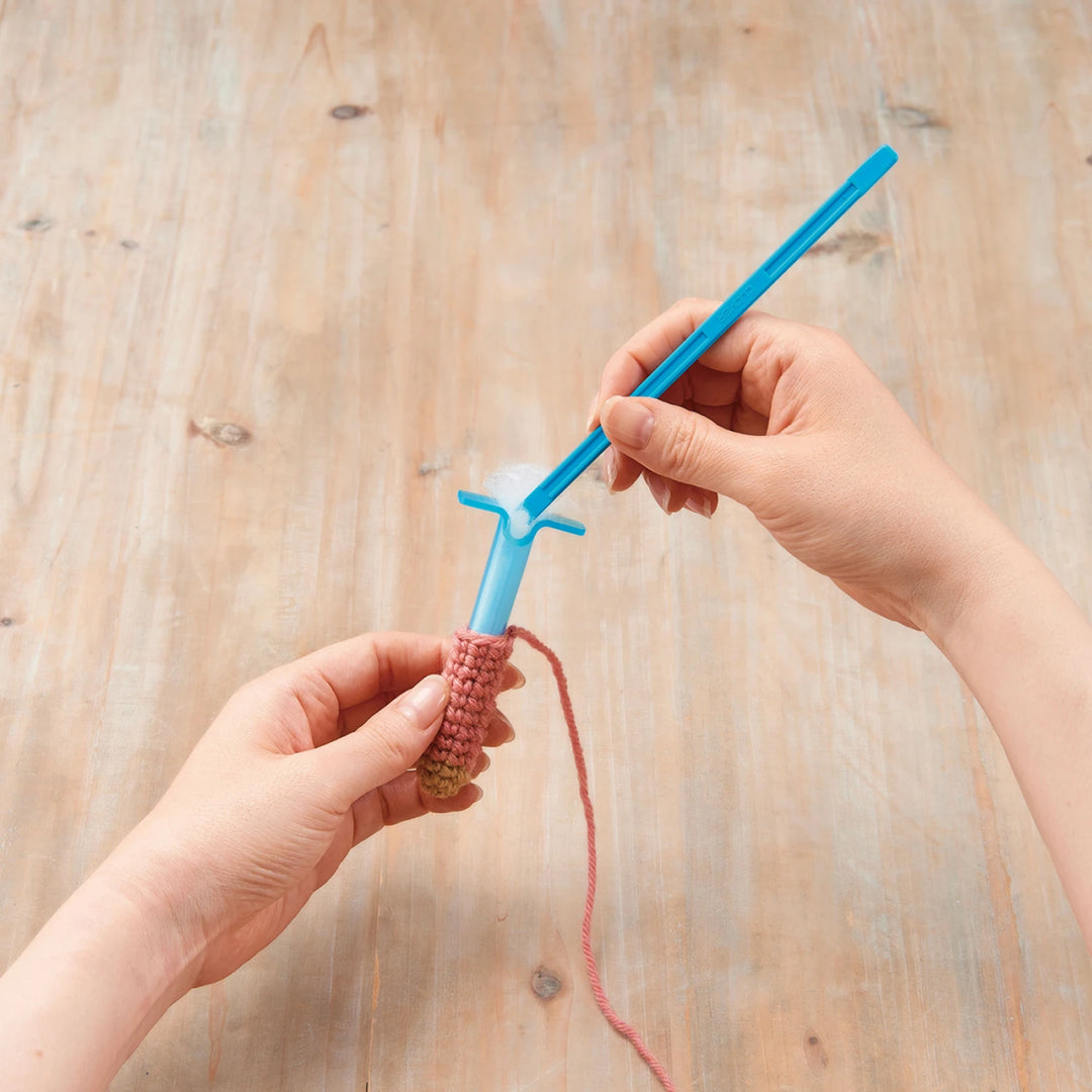 Hands using a blue Tube & Plunger Style Stuffing Tool to fill a small crochet or knit project with pink yarn on a wooden surface in a modern needlework craft setting.