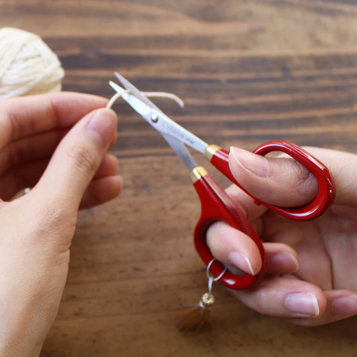Hand holding soft white thread being cut with Japanese Artisan Lacquered Handle Embroidery Scissors with red lacquered handles on wooden surface, suitable for embroidery and needlework crafts.
