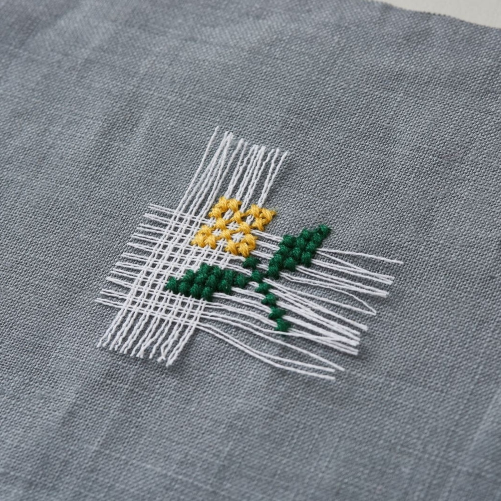 Close-up of delicate embroidery on gray fabric using white Cross Stitch Waste Canvas to create a small yellow flower with green stem, showcasing modern needlework technique.
