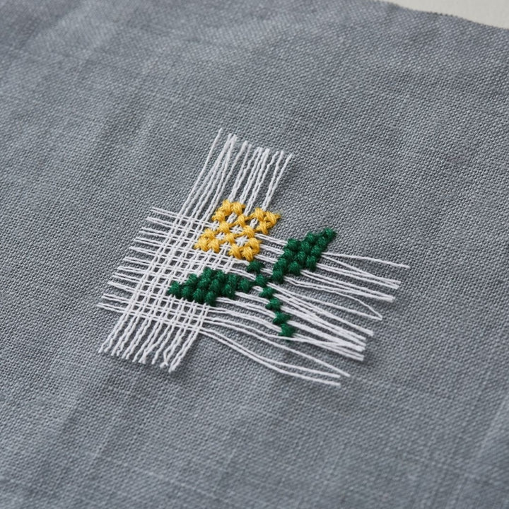 Close-up of delicate embroidery on gray fabric using white Cross Stitch Waste Canvas to create a small yellow flower with green stem, showcasing modern needlework technique.