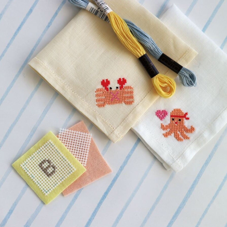 Cross Stitch Waste Canvas with small completed cross stitch designs of a crab and octopus on fabric, accompanied by embroidery floss in yellow and blue on a striped background.