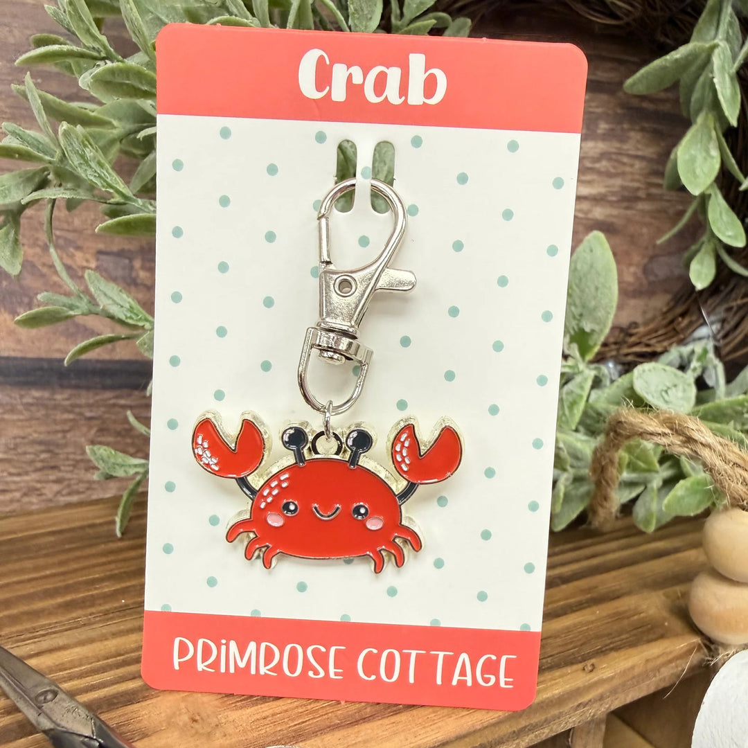 Red crab keychain on a polka dot card with 'Primrose Cottage' branding, placed on a wooden surface with plants in the background.