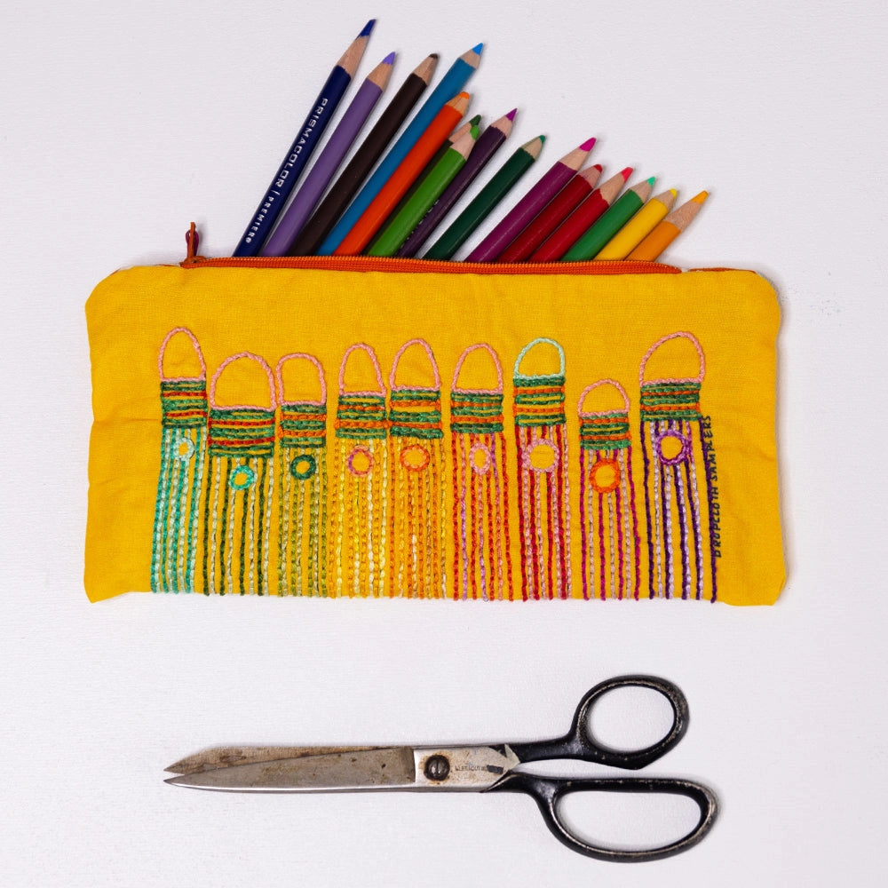 Bright yellow fabric pouch featuring colorful pencil designs embroidered with vibrant threads, holding colored pencils above vintage scissors, showcasing the Pencils Embroidery Sampler.