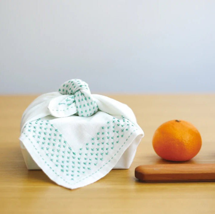 Half-Yard 5mm Straight Line Grid Sashiko Sampler cloth with delicate green sashiko stitching displayed wrapped around a small box on a wooden surface alongside a fresh orange.