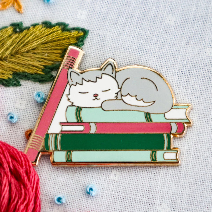 Pin with a design of a cat sleeping on books, surrounded by embroidery materials.