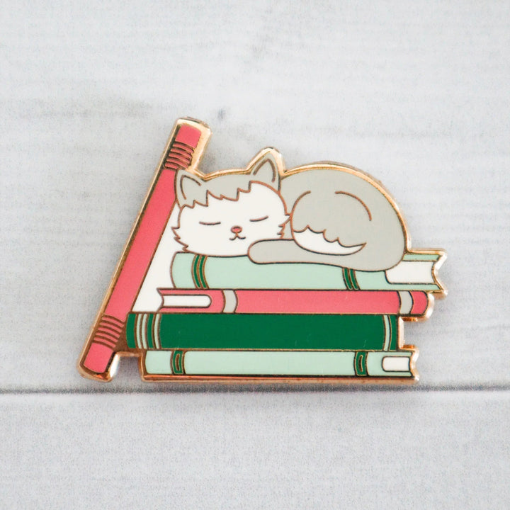 Enamel pin of a cat sleeping on books with a pencil next to it on a light gray background