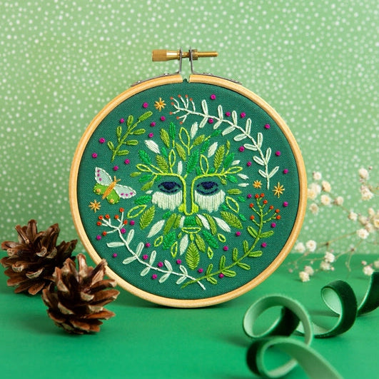 A circular embroidery hoop displaying a green tree-of-life face surrounded by leaves, flowers, and a butterfly, styled on a green surface with pinecones and ribbon against a green patterned backdrop.