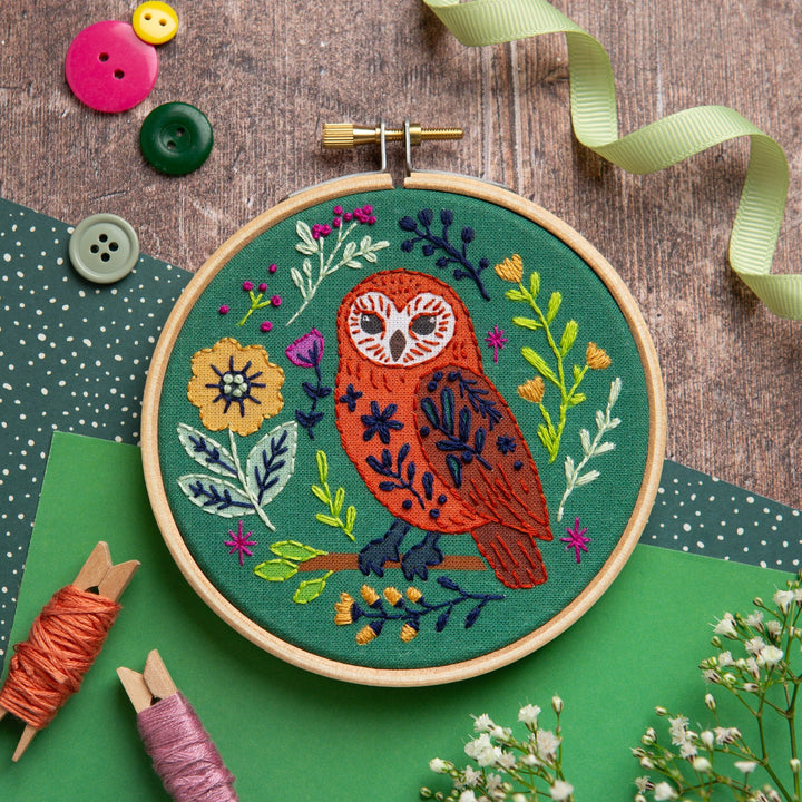 Embroidery hoop with owl design on a wooden surface with buttons and thread spools.