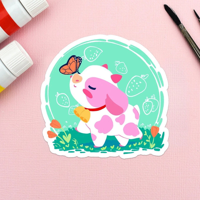 A die-cut sticker featuring a cute cartoon pig in a mint green circular frame, styled on a pink textured background with paint tubes and paintbrushes.