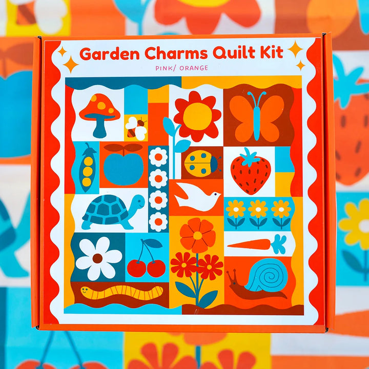 Garden Charms Whole Cloth Quilt Kit featuring colorful, whimsical garden-themed illustrations including flowers, a butterfly, apple, turtle, snail, and other nature-inspired designs in vibrant pink and orange tones.