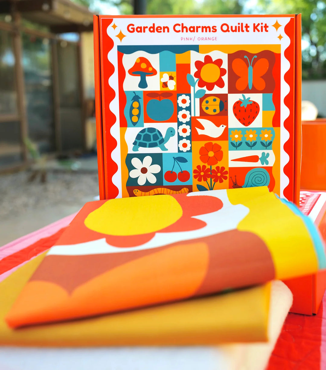 Garden Charms Whole Cloth Quilt Kit featuring vibrant pink and orange fabric with playful nature-inspired designs including flowers, animals, and fruit, perfect for modern needlework projects.