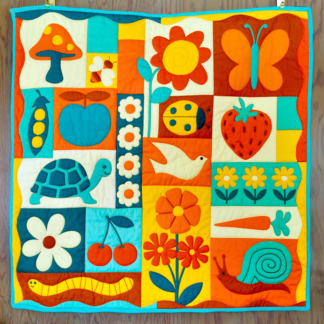 Garden Charms Whole Cloth Quilt Kit featuring vibrant hand-stitched designs of animals, flowers, fruits, and insects in bright blue, orange, yellow, and red tones on quilted fabric.