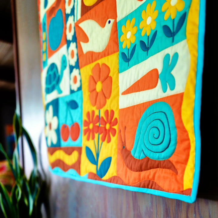Close-up of colorful garden-themed quilt featuring flowers, a carrot, a snail, and a bird, made with vibrant blue and orange fabric, showcasing the Garden Charms Whole Cloth Quilt Kit.
