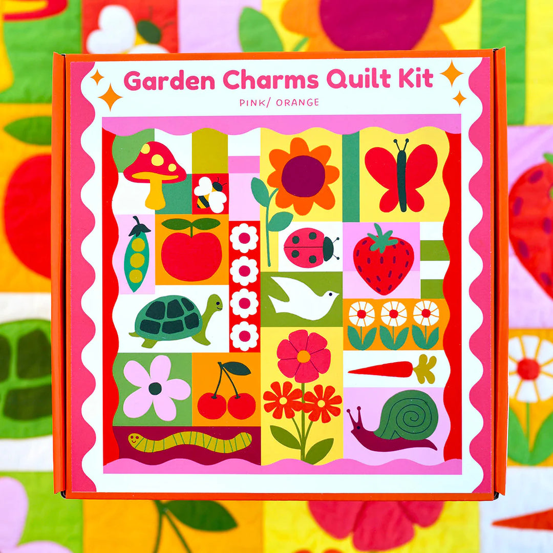 Garden Charms Whole Cloth Quilt Kit box featuring colorful illustrations of a mushroom, flower, butterfly, turtle, strawberry, cherries, bird, ladybug, carrot, snail, and other garden-themed designs in pink, orange, red, and green tones.