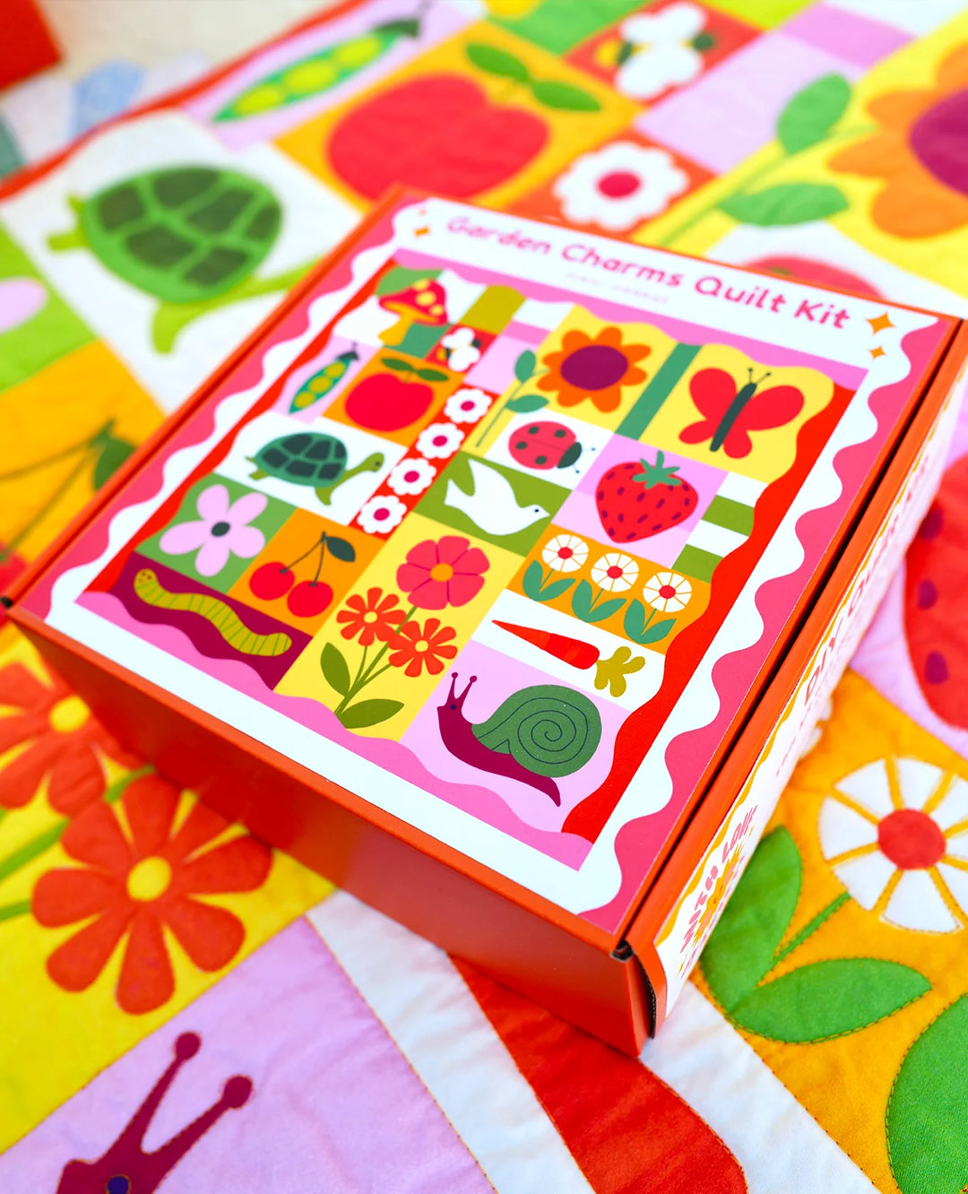 Brightly colored Garden Charms Whole Cloth Quilt Kit box featuring playful garden-themed illustrations like flowers, a snail, turtle, butterfly, and strawberry on a vibrant quilt background.