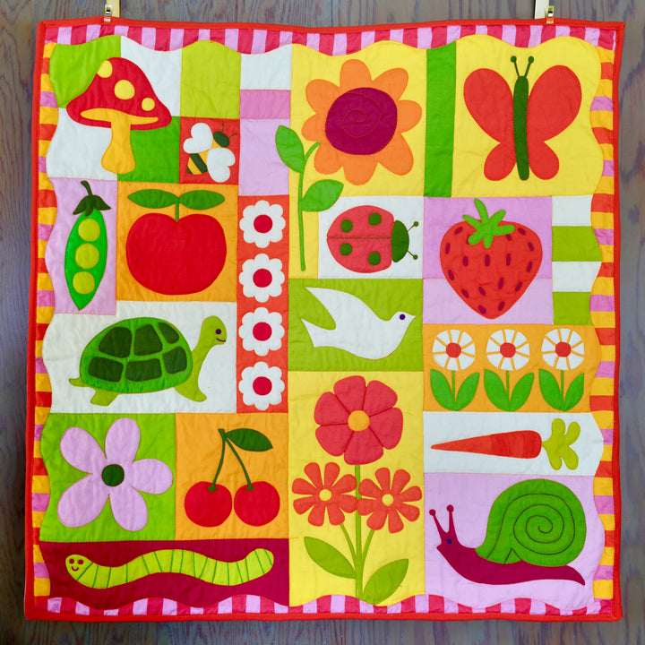 Garden Charms Whole Cloth Quilt Kit featuring bright pink, green, yellow, and orange modern needlework designs including a butterfly, turtle, flowers, and fruits on quilted fabric.