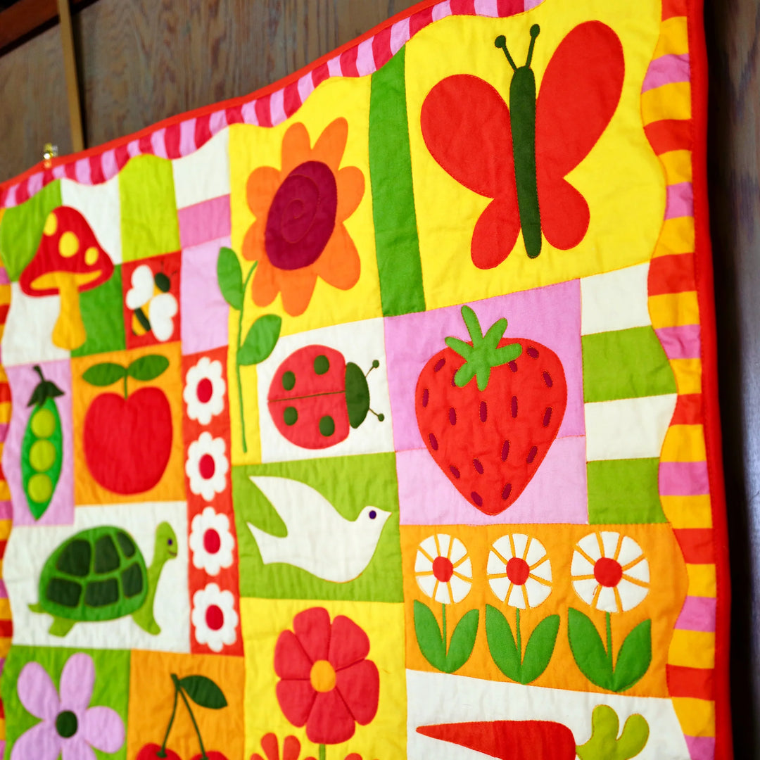 Close-up of a colorful quilt featuring appliqué designs of a butterfly, strawberry, ladybug, turtle, and flowers in pink, green, red, and yellow hues, part of the Garden Charms Whole Cloth Quilt Kit.
