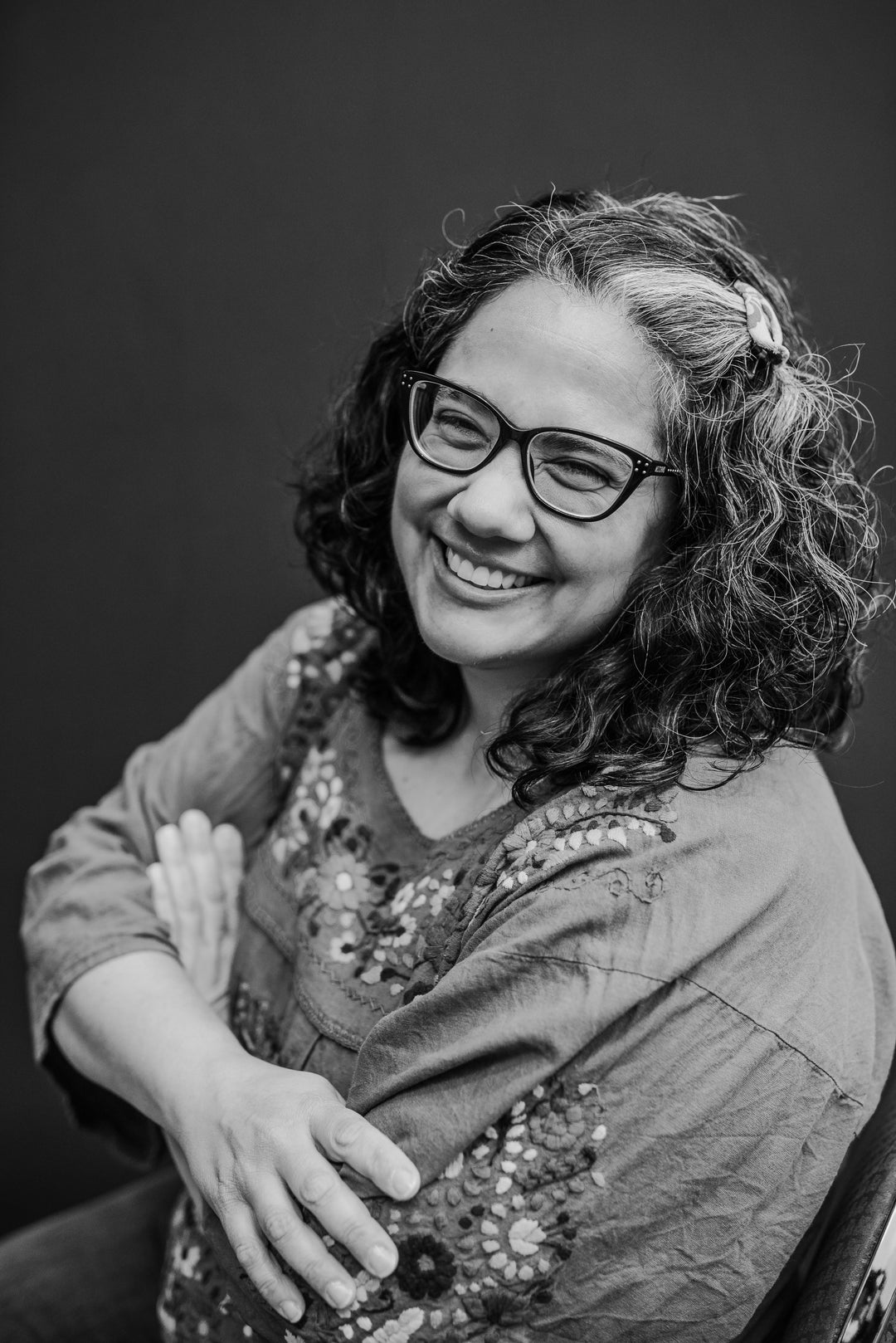 Black and white portrait of a smiling woman with curly hair and glasses wearing an embroidered top, seated with arms crossed against a plain dark background.