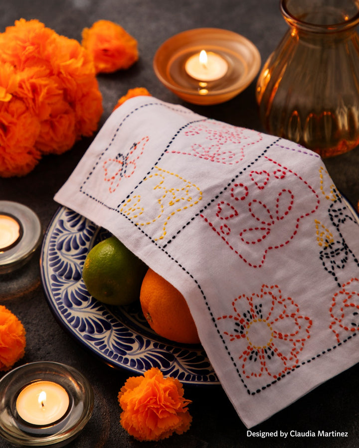 A Day of the Dead themed Sashiko sampler draped over a plate of fruit on an Ofrenda surrounded by candles and cempasuchil