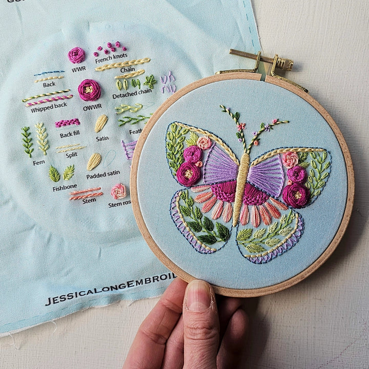 Hand holding an embroidery hoop with a colorful butterfly stitched using various embroidery floss techniques on blue fabric, next to a detailed stitch guide, for the Beginner Butterfly Sampler Hand Embroidery Kit.