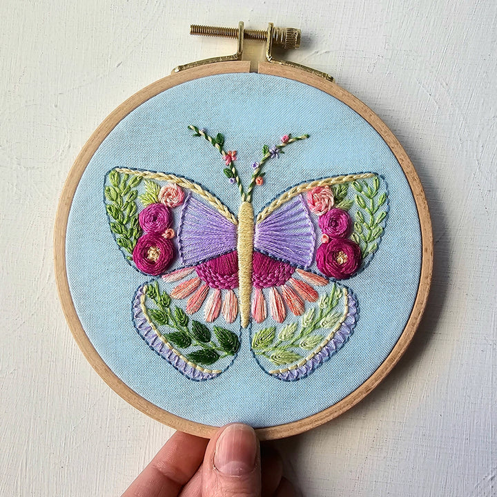 Hand embroidery hoop featuring a colorful butterfly stitched with vibrant embroidery floss on light blue fabric, showcasing the Beginner Butterfly Sampler Hand Embroidery Kit.