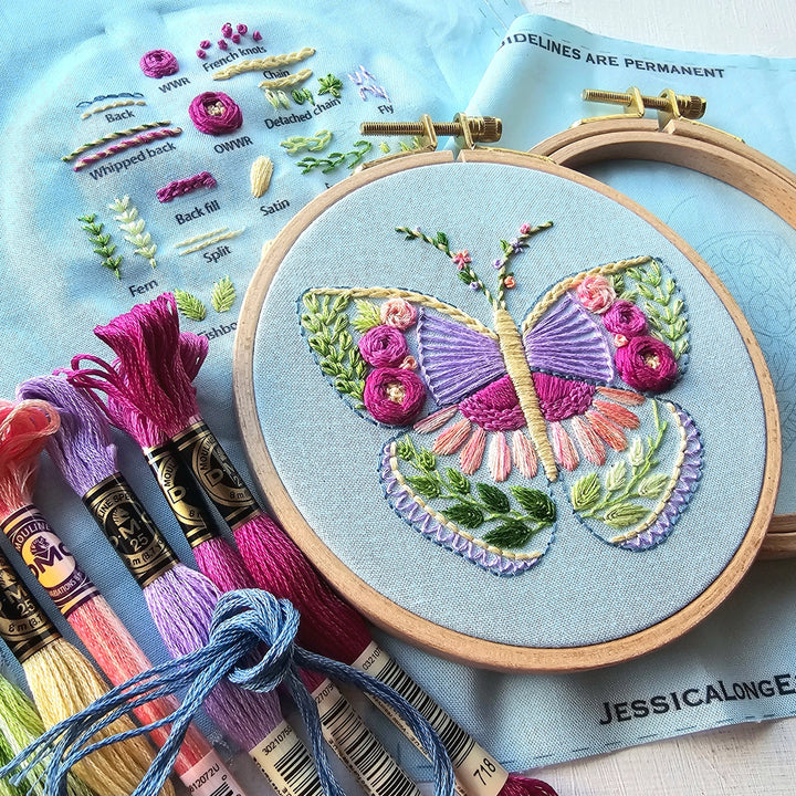 Beginner Butterfly Sampler Hand Embroidery Kit featuring a colorful embroidered butterfly with floral details on aida cloth, surrounded by embroidery floss in shades of pink, purple, and blue.