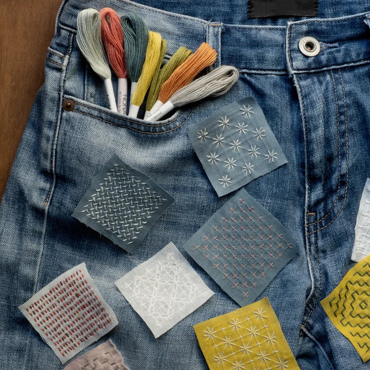 A close-up of a blue denim jeans pocket holding several embroidered fabric swatches and a set of multicolored embroidery floss threads, arranged on a wooden surface.