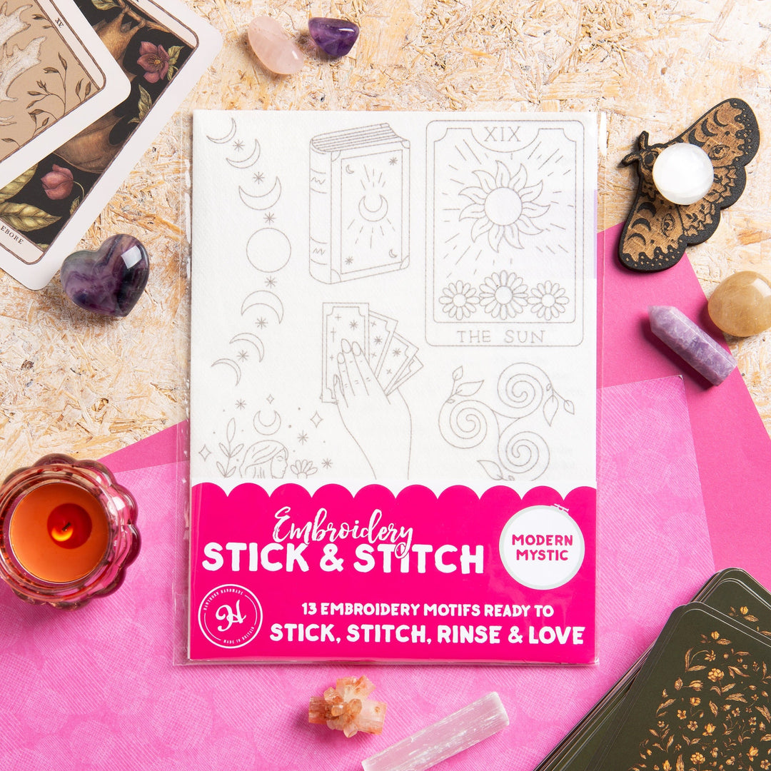 Embroidery kit packaging with pink border on a textured surface with tarot cards and decorative items.