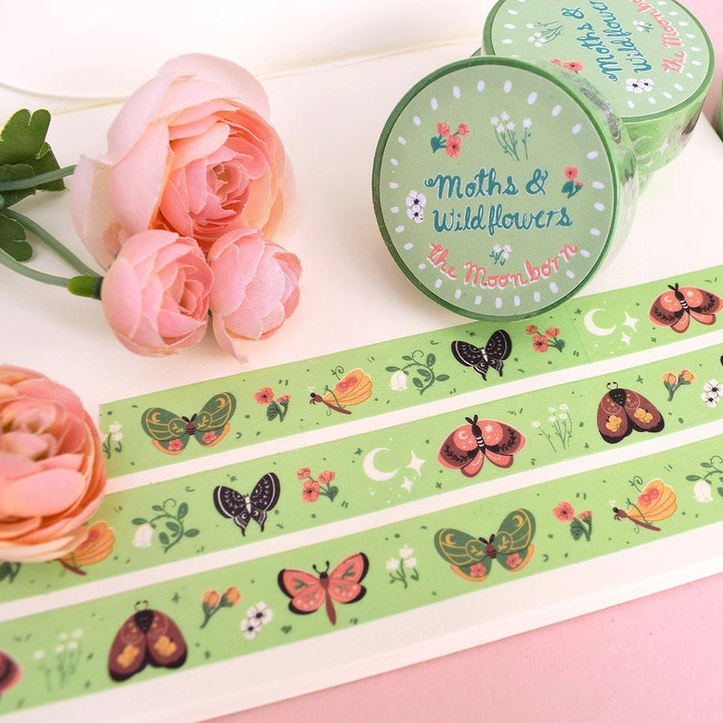 Green washi tape with a whimsical moth and wildflower pattern, shown unrolled on a white notebook alongside three matching rolls. Pink roses are placed on the left, set against a soft pink background.