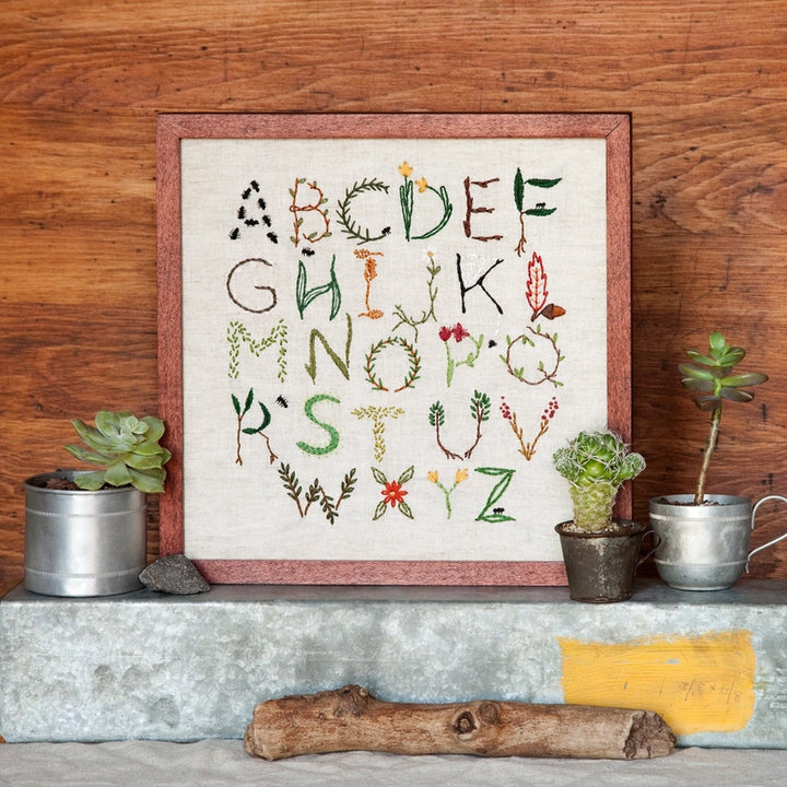 Nature Inspired Alphabet Embroidery Sampler featuring colorful hand-stitched letters with botanical motifs, displayed in a wooden frame with small potted plants on a rustic surface.