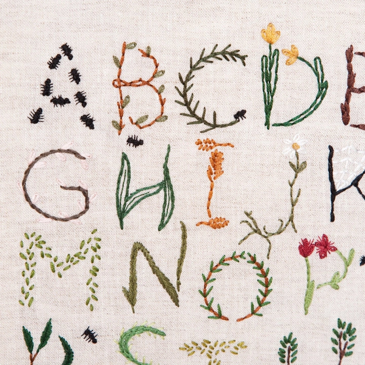 Close-up of embroidered alphabet sampler featuring nature-inspired letters with floral and leaf motifs in various colors on fabric, showcasing the Nature Inspired Alphabet Embroidery Sampler.