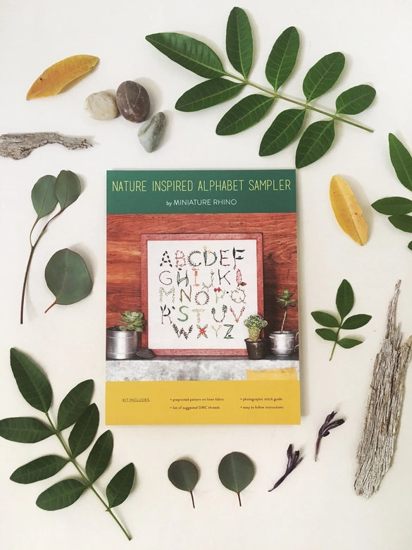 Nature Inspired Alphabet Embroidery Sampler kit cover featuring delicate botanical-themed alphabet stitching, surrounded by green leaves, twigs, and natural stones.