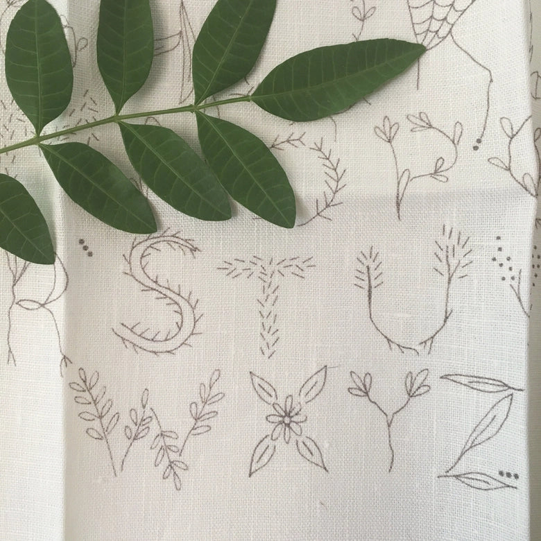 Close-up of detailed hand embroidery featuring natural botanical motifs forming letters on light fabric, part of the Nature Inspired Alphabet Embroidery Sampler displayed with green leaves.