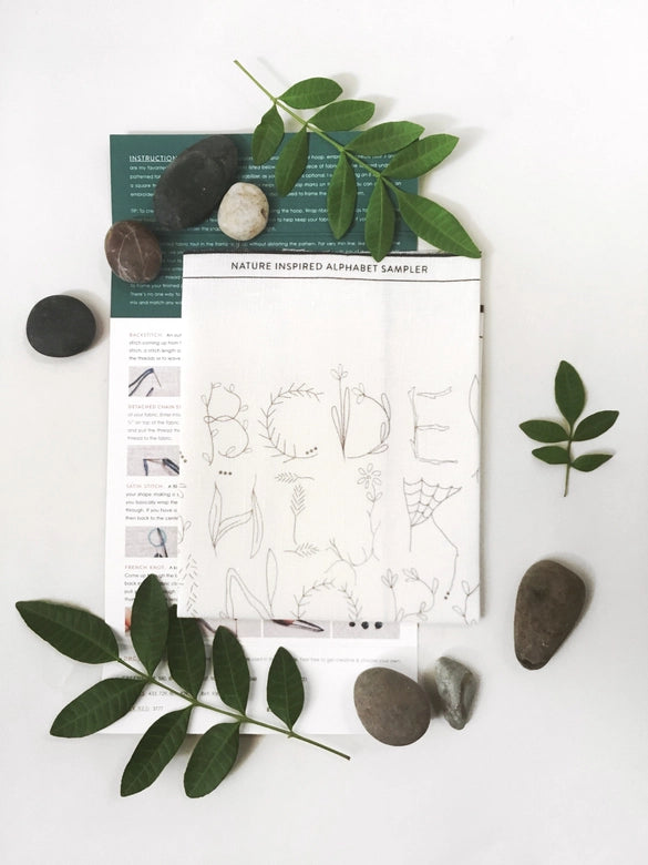 Nature Inspired Alphabet Embroidery Sampler fabric featuring delicate botanical letter designs, displayed with smooth stones and green leaves for a natural craft store presentation.